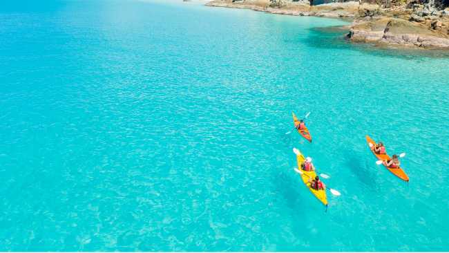 Kayakers paddle turquoise waters off a forested island in the Whitsundays, Queensland.